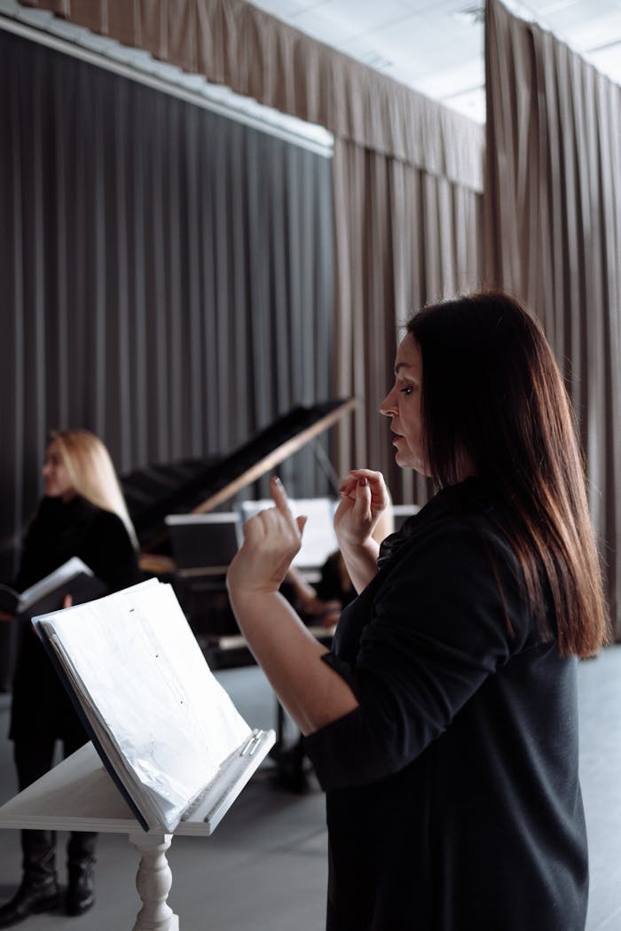 A female conductor gestures while leading an orchestra rehearsal indoors.