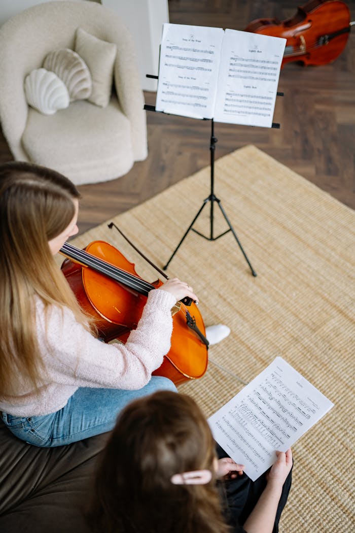 A young girl is learning to play the cello with her teacher at home. Musical notes are visible.